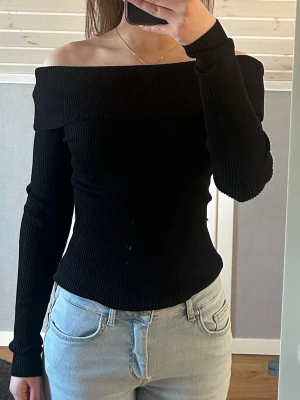 Svart ribbad offshoulder topp - Snygg svart ribbad offshoulder topp med lång ärm och figurnära passform. Toppen har en bred kant över axlarna som ger en snygg siluett och är perfekt för dig som gillar stilrena plagg med det lilla extra. Materialet är stretchigt och bekvämt. Använd två gånger