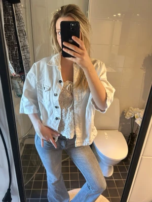 Vit jeansjacka med korta ärmar - Säljer en vit jeansjacka med korta ärmar och klassisk krage. Jackan har två bröstfickor med knappar och en avslappnad, boxig passform. Perfekt att slänga över en topp för en cool och fräsch look.