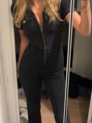 Svart jumpsuit med dragkedja - Snygg svart jumpsuit i denim-look med korta ärmar, krage och dragkedja framtill. Två bröstfickor med knappar och markerad midja ger en cool och trendig siluett. Perfekt till sneakers eller boots för en edgy stil.