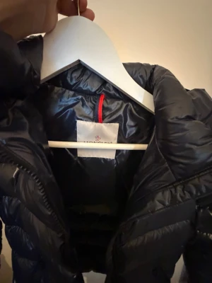 Svart dunjacka från Moncler - Snygg svart dunjacka från Moncler med huva och quiltad design. Jackan har en glansig finish och dragkedja framtill. Perfekt för kalla vinterdagar och ger en stilren look. Klassisk Moncler-logga på insidan.