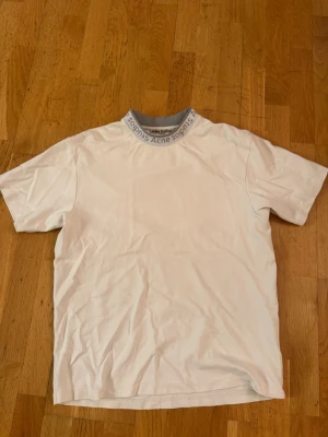 Vit t-shirt från Acne Studios - Vit t-shirt från Acne Studios med grå ribbad krage där det står 'Acne Studios' runtom. Klassisk passform och korta ärmar. Perfekt till jeans eller shorts för en clean och stilren look.