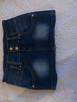 Blå jeanskjol med fickor och knappar - Säljer en blå jeanskjol med snygga slitningar och kontrastsömmar. Kjolen har flera fickor både fram och bak samt dekorativa knappar framtill. Perfekt för dig som gillar en casual och cool stil.