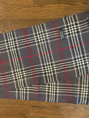 Grå Burberry Halsduk - Klassisk halsduk i grått med vitt, svart och rött rutmönster från Burberry. Tillverkad i mjuk ull som känns lyxig och varm. 