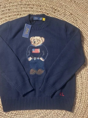 Mörkblå stickad tröja Polo Bear - Mörkblå stickad tröja från Polo Ralph Lauren med ikonisk Polo Bear-motiv på bröstet. Tröjan har rund halsringning, ribbade muddar och RL-broderi vid nederkanten. Materialet är mjukt och bekvämt, perfekt för en avslappnad men snygg look. Tröjan är helt ny och storlek m hör av er vid frågor eller funderingar priset går att diskutera!!