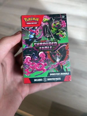 Pokémon Shrouded Fable Booster Bundle - Pokémon Scarlet & Violet Shrouded Fable Booster Bundle med 6 booster packs. Förpackningen är färgglad med gröna, rosa och svarta detaljer och visar flera Pokémon-karaktärer. Perfekt för samlare och fans av Pokémon TCG.