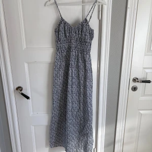 Långklänning med blåvitt mönster - Säljer en långklänning med smala axelband och ett blåvitt grafiskt mönster. Klänningen har v-ringad hals och resår i midjan för en bekväm passform. Perfekt för varma dagar och sommarens alla äventyr.