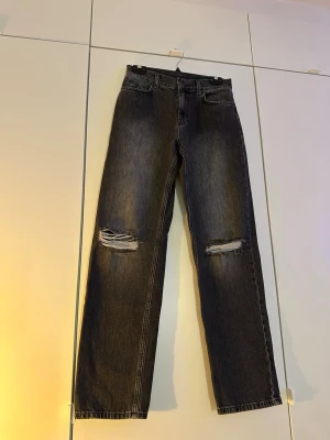 Svart gråa raka jeans med slitningar - Säljer ett par svarta gråa raka jeans med slitna hål över båda knäna. Jeansen har klassisk femficksmodell och hög midja. Materialet är denim i bomull och färgen är tvättad svart för en cool vintage-look.