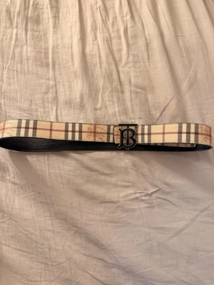 Burberry bälte med klassiskt rutmönster - Snyggt bälte från Burberry med det ikoniska beigea och bruna rutmönstret på ena sidan och svart på andra sidan. Spännet har en metallisk logga i silver. Perfekt accessoar för att lyfta din outfit.