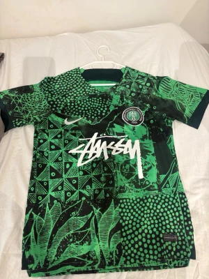 Grön Nigeria fotbollströja Nike M - Säljer en grön Nigeria fotbollströja från Nike med Dri-Fit material. Tröjan har ett unikt mönster i olika gröna nyanser, svart och vitt, samt Stüssy-logga framtill. Korta ärmar och rund hals. Perfekt för dig som gillar streetwear och sport.
