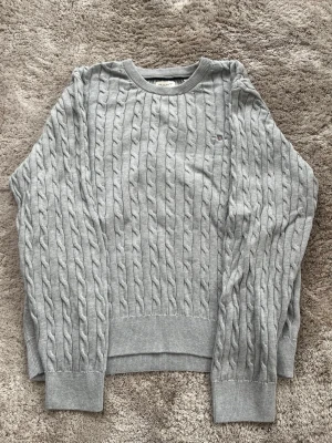 Ljusgrå kabelstickad tröja Tommy Hilfiger - Säljer en ljusgrå kabelstickad tröja från Tommy Hilfiger med rund halsringning och diskret logga broderad på bröstet. Tröjan har ribbade muddar vid ärmslut och nederkant, och är tillverkad i mjukt material som passar perfekt till alla tillfällen. OBS! Barnstorlek 176