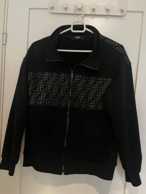 Svart zip från Fendi - Snygg svart zip från Fendi med hel dragkedja och ikoniskt FF-mönster i grått över bröstet och axlarna. Tröjan har två fickor och en hög krage. Storlek S men passar även XS. Pris kan diskuteras vid snabb affär!