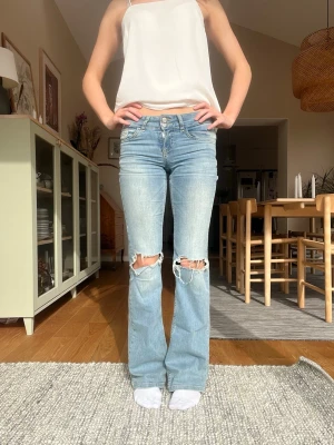 Ljusblå bootcut jeans med slitningar - Säljer ett par ljusblå bootcut jeans med slitna hål över knäna och klassiska fem fickor. Jeansen har en snygg tvätt och är tillverkade i denim med lite stretch för skön passform. Perfekta för en avslappnad och trendig look.