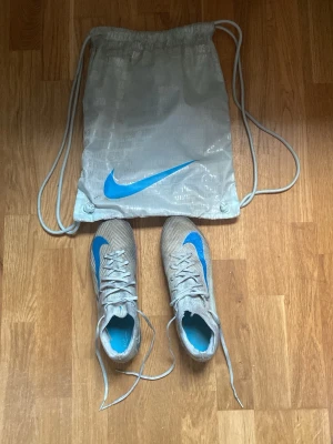 Nike Mercurial air Elite nypris 3400kr - Säljer ett par Nike Mercurial fotbollsskor i silver med blå detaljer och snörning. Skorna har en strömlinjeformad design och tillhörande gympåse med blå Nike-logga. Yttermaterialet är syntetiskt och sulan har fasta dobbar för spel på gräs och konstgräs. Ena skon har en väldigt liten skada som man ser på sista bilden men inget som stör.