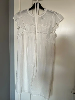 Vit kortklänning med volangärmar - Söt vit kortklänning med prickigt mönster, volangärmar och knytband i nacken. Klänningen har en romantisk känsla med fina spetsdetaljer upptill och är tillverkad i ett lätt, luftigt material. Perfekt för varma dagar eller sommarens alla tillfällen.