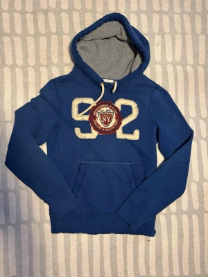 Blå hoodie Abercrombie & Fitch - Blå hoodie från Abercrombie & Fitch med stor vit 92-print och NY-märke framtill. Mjuk insida, ribbade muddar och känguruficka. Dragsko i huvan, hör av dig vid funderingar 