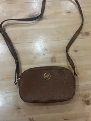 Brun axelväska från Michael Kors - Snygg brun axelväska från Michael Kors i äkta läder. Väskan har ett stilrent guldigt MK-emblem framtill och justerbar axelrem. Perfekt storlek för det viktigaste och enkel att matcha med olika outfits.