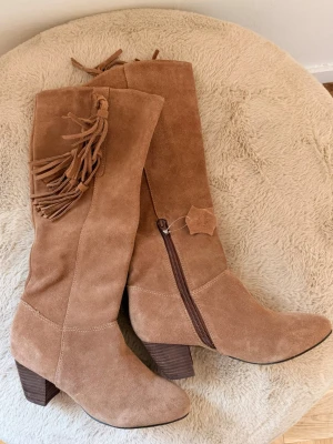 Bruna mocka cowboy boots med fransar Boho Chic - Snygga cowboy boots i ljusbrun mocka med dekorativa fransar på skaftet och snedklack i trälook. Dragkedja på insidan för enkel påtagning och spetsig tå ger bootsen en klassisk westernstil. Perfekta för dig som vill sticka ut med en trendig western vibe och festivaler