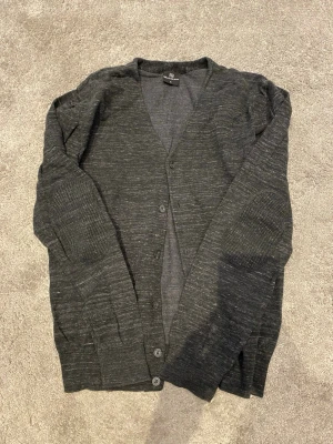 Mörkgrå kofta från Mille Milano - Stilren mörkgrå kofta från Mille Milano med knäppning framtill och långa ärmar. Tillverkad i mjukt, tunt stickat material som passar perfekt för lager på lager. Klassisk v-ringad design och ribbade muddar vid ärmslut och nederkant.