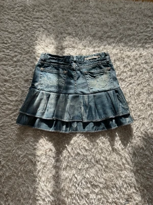 Blå volangkjol i jeans med detaljer - En jeanskjol omsydd från ett par jeans med coola detaljer.  Midjemått tvärsöver:36cm Längd: 36cm