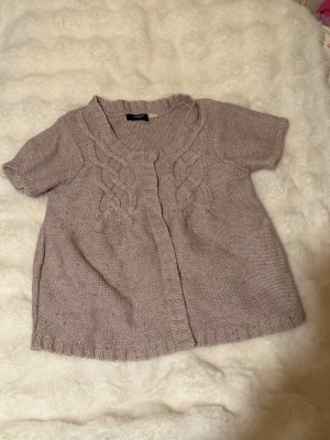 Ljuslila/beige stickad kofta Redoute - Kortärmad stickad kofta i ljuslila/beige från Redoute Création. Koftan har ett snyggt flätmönster framtill, rundad halsringning och ribbade kanter. Perfekt att slänga över en topp för extra stilpoäng.