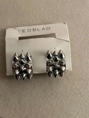 Silvriga nitar örhängen från Edblad - Coola örhängen från Edblad i blankt silverfärgat metallmaterial. De har en rektangulär form och är dekorerade med flera små, spetsiga nitar som ger en edgy och modern look. Perfekta för dig som gillar statement-smycken och vill sticka ut. LÅNAD BILD!