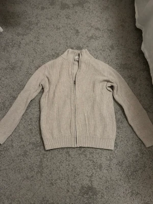 Beige stickad ziptröja med hög krage - Säljer en beige stickad ziptröja med hög krage och ribbade muddar. Tröjan har en rak passform och dragkedja framtill. Perfekt för lager på lager och enkel att matcha med olika outfits. Storlek 152 men passar även dig som har en lite större storlek.
