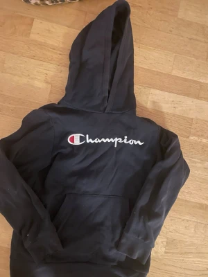 Svart Champion hoodie med logga - Säljer en svart hoodie från Champion med stor broderad logga på bröstet. Klassisk känguruficka framtill och stor huva. Tillverkad i mjukt bomullsmaterial som är skönt att ha på sig. Perfekt för dig som gillar streetwear och sportig stil.