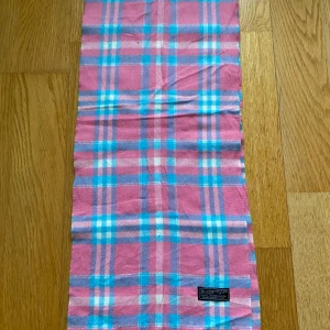 Rosa-rutig halsduk Burberry - Säljer en rosa halsduk från Burberry med klassiskt rutmönster i vitt, blått och ljusrosa. Halsduken är rektangulär och har en mjuk känsla, perfekt för att addera färg till din outfit. Märkeslappen sitter fast i ena hörnet.