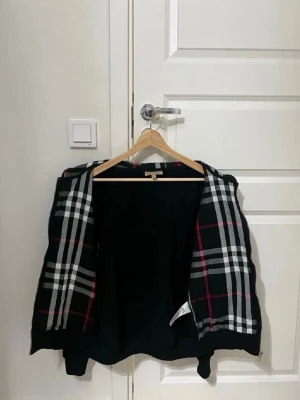 Svart hoodie från Burberry med rutigt foder - Snygg svart hoodie från Burberry med klassiskt rutigt foder i svart, vitt och rött. Dragkedja framtill och broderad Burberry-logga på bröstet. Perfekt för dig som vill ha en hoodie med exklusiv känsla och stilrena detaljer.