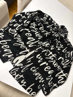 Svart overshirt med vit text från Zara - Snygg svart overshirt från Zara med vit grafisk text över hela plagget. Overshirten har klassisk krage, knäppning framtill och två bröstfickor med lock. Materialet ser ut att vara mjukt och lite tjockare, perfekt för lager på lager. Riktigt trendig och unik stil!