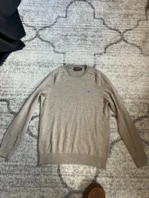 Beige stickad tröja från Morris - Snygg beige stickad tröja från Morris med rund halsringning och diskret blå logga broderad på bröstet. Tröjan har långa ärmar och ribbade muddar vid ärmslut och nederkant. Perfekt för en stilren och avslappnad look.