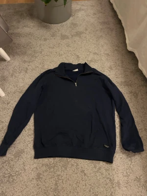 Mörkblå halvzip tröja - Mörkblå långärmad tröja med halvzip och hög krage. Tröjan har ribbade muddar vid ärmslut och nederkant samt en diskret etikett nertill. Perfekt för en avslappnad och stilren look.