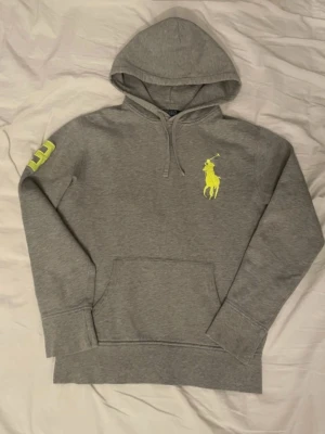 Ralph Lauren hoodie - Snygg grå hoodie från Polo Ralph Lauren med stor gul logga på bröstet och siffran 3 i gult på ärmen. Tillverkad i en mjuk bomullsblandning med huva och känguruficka framtill. Perfekt för en avslappnad och trendig stil.