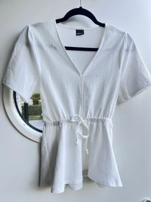 Vit peplumtopp med knytband i midjan, Gina Tricot - stl S - Vit blus/peplumtopp från Gina Tricot med v-ringning. Knytband i midjan för en perfekt passform och har ett strukturerat material. Aldrig använd.