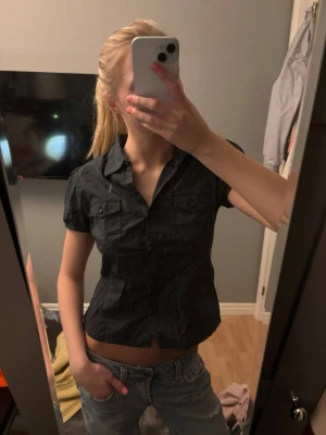 Svart kortärmad skjorta med knappar - Snygg svart skjorta med korta ärmar och knappar framtill. Skjortan har två bröstfickor med lock och en figurnära passform. Köpt i london, använd några gånger.  Passar nån som bär S. 