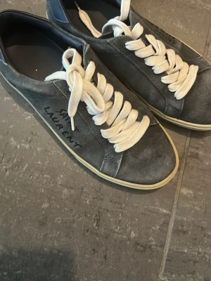 Grå Saint Laurent sneakers - Snygga grå sneakers från Saint Laurent med vita, breda skosnören och diskret logga på sidan. Skorna har en klassisk låg profil och ovandel i mocka som ger en stilren och trendig look. Perfekta för dig som gillar exklusiva märken och enkel design.