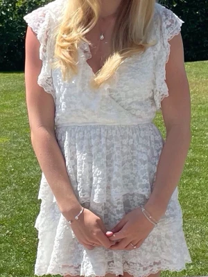 Vit spetsklänning med volang - Söt vit kortklänning i spets med omlott upptill och volangdetaljer. Klänningen har v-ringning och är ärmlös med breda axelband. V-ringad öppen rygg med ett smalt knyttband. Perfekt för sommarens alla tillfällen.