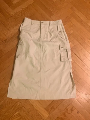 Beige långkjol med cargofickor - Stilren beige långkjol med cargofickor på sidorna och bälteshällor i midjan. Kjolen har knapp- och dragkedjestängning framtill samt slits i sidan för extra rörelsefrihet. Perfekt för en avslappnad men trendig look.Skriv gärna för mått! 💕