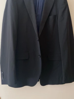  svart kavaj - Stilren svart kavaj med två knappar framtill, Slim fit helfoder i mörkblått. Perfekt för en dressad look. Bredd mellan Axlarna 41cm, längd 72cm,  midja ungefär 80c. Längd ärmar,66vm