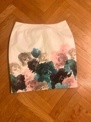 Vit blommig kortkjol med dragkedja - Säljer en vit kortkjol med stora blommor i pastellfärger som rosa, blågrön och beige. Kjolen har en smal passform och dragkedja bak. Perfekt för dig som vill ha en feminin och trendig look.Skriv gärna för mått! 💕