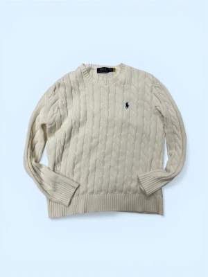 Vit kabelstickad tröja Polo Ralph Lauren - Kabelstickad vit tröja från Polo Ralph Lauren med rund halsringning och ribbade muddar. Klassisk marinblå logga broderad på bröstet. Tröjan har lång ärm och är gjord i mjukt material, perfekt för kyliga dagar.