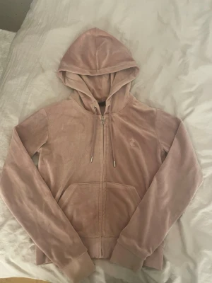 Rosa tröja från Juicy Couture - Säljer en klassisk rosa velourhoodie från Juicy Couture med dragkedja framtill och fickor. Hoodien har en broderad JC-logga på bröstet, justerbar huva med snören och ribbade muddar. Perfekt för en chill och trendig look.