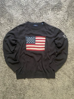 Svart stickad tröja från Gant - Stickad svart tröja från Gant med amerikansk flagga i rött, vitt och blått på bröstet. Rund halsringning och ribbade muddar. Diskret text på ärmen. Perfekt för dig som gillar klassisk college-stil.