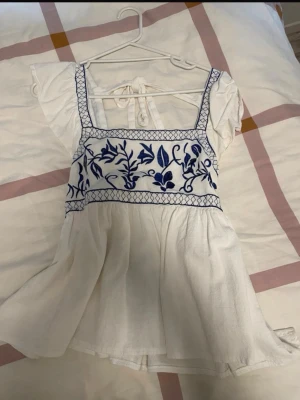 Vit blommig kort klänning med knyt - Söt vit kort klänning med blå broderade blommor över bysten och rutiga detaljer vid axelbanden. Klänningen har korta ärmar, knyt i ryggen och är gjord i ett lätt bomullstyg. Perfekt för sommarens varma dagar.