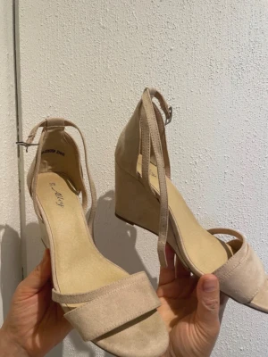 Beige kilklackade sandaler Ally - Snygga beige sandaler från Ally med kilklack och öppen tå. Skorna har en smal rem över tårna och justerbar ankelrem med spänne. Tillverkade i mockaimitation för en trendig och stilren look.