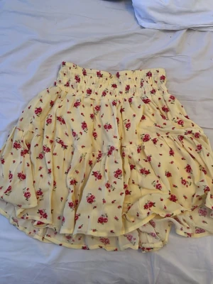 Gul blommig volangkjol - Söt gul kortkjol med små röda blommor och volangdetaljer. Kjolen har bred resår i midjan för skön passform och ett luftigt, flowy tyg som passar perfekt till varma dagar. Perfekt för dig som gillar romantisk och somrig stil.