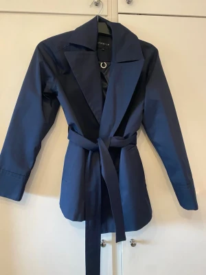 Mörkblå trenchcoat från Stockh LM - Stilren mörkblå trenchcoat från Stockh LM med klassisk krage, knytband i midjan och raka ärmar. Jackan har en minimalistisk design och är tillverkad i ett slätt tyg med lätt glans. Perfekt för dig som gillar en clean och modern look. Aldrig använd 