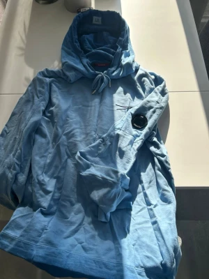 Blå hoodie från C.P. Company - Snygg ljusblå hoodie från C.P. Company med huva och dragsko. Ikonisk linsdetalj på ärmen och etiketten 'RE-COLOUR' i nacken. Tillverkad i mjukt bomullsmaterial, perfekt för en avslappnad streetwear-look. 