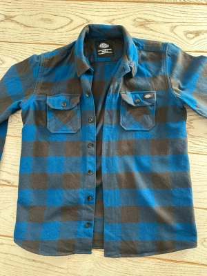 Blå och svart rutig skjorta Dickies - Säljer en blå och svart rutig skjorta från Dickies med klassisk krage och två bröstfickor med knappar. Skjortan har långa ärmar och knäppning framtill. Perfekt för en avslappnad och cool stil.