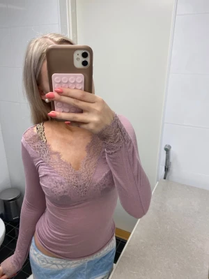 Ljuslila långärmad topp med spets - Säljer en ljuslila långärmad topp med v-ringning och fina spetsdetaljer runt halsen och ärmsluten. Toppens passform är tight och den är gjord i ett mjukt material som känns skönt mot huden. Perfekt för dig som gillar romantiska detaljer och en feminin look.